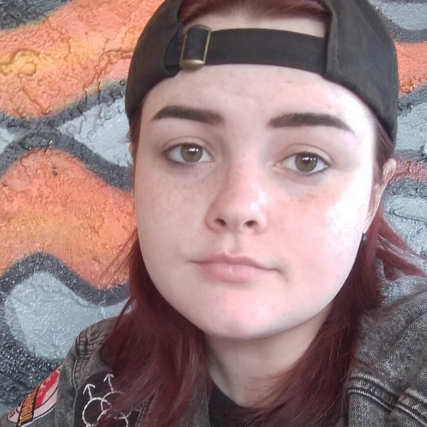 A selfie of me with red hair, a black ball cap, green eyes with smudge black eyeliner on the bottom lid and a jean jacket in front of a flamed mural wall